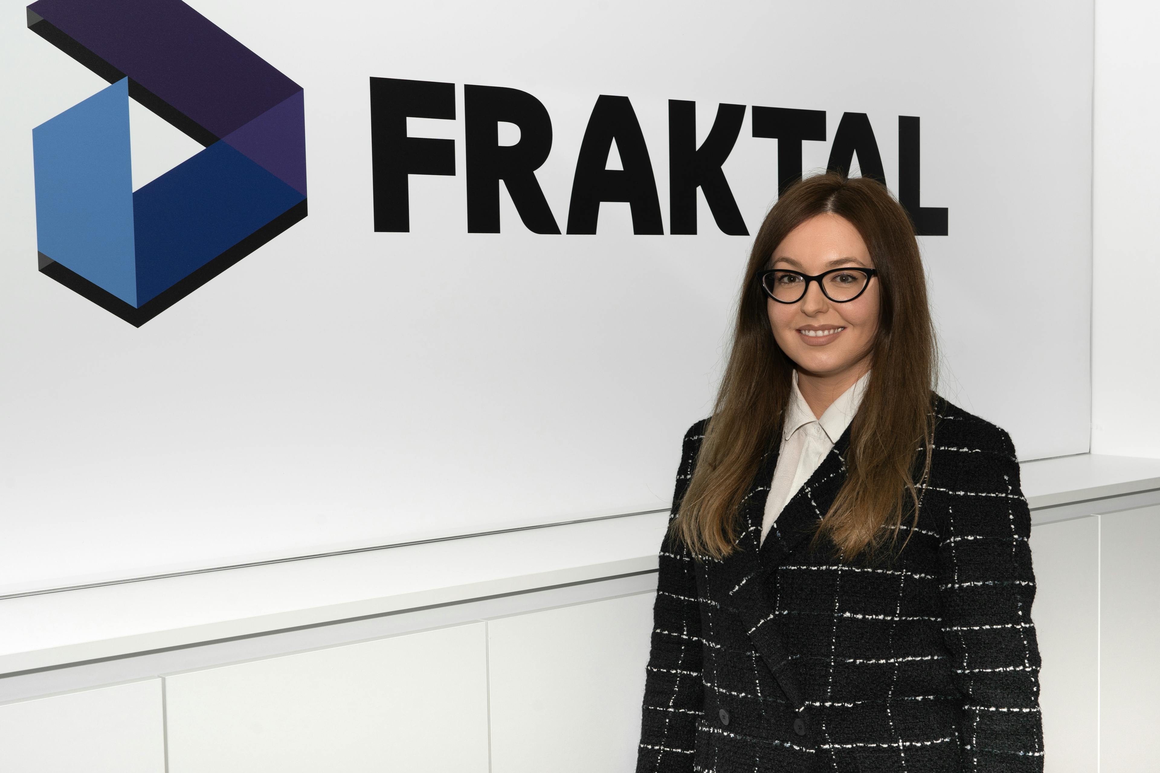 Mia Mikulic from the administration team at Fraktal Development, standing confidently with a friendly expression. She wears stylish glasses, a chic black and white tweed jacket over a crisp white shirt, projecting professionalism and approachability. The bold 'FRAKTAL' logo in the background affirms the corporate identity.