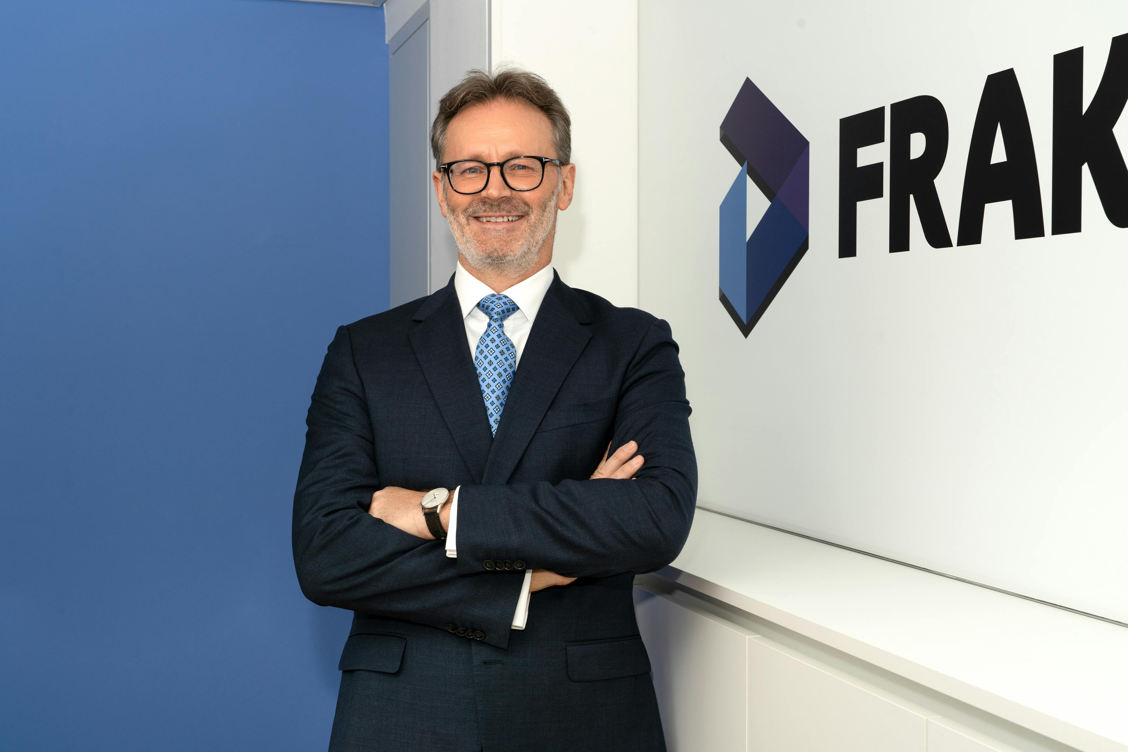 Portrait of Anthony Pink, a partner at Fraktal Development, poised confidently with arms folded in front of a panel displaying the 'FRAKTAL' logo. He is dressed in a professional navy blue suit, patterned blue tie, and glasses, offering a friendly smile in a well-lit office setting that reflects the company's branding colors.
