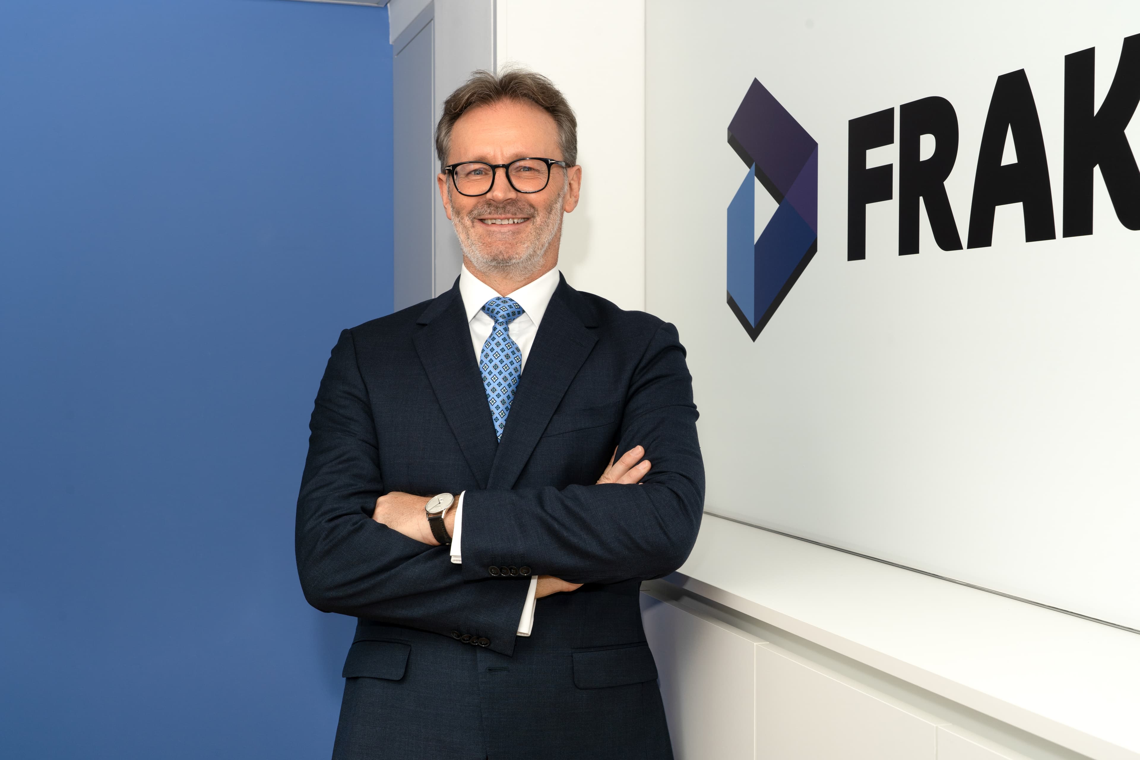 Portrait of Anthony Pink, the director at Fraktal Development, poised confidently with arms folded in front of a panel displaying the 'FRAKTAL' logo. He is dressed in a professional navy blue suit, patterned blue tie, and glasses, offering a friendly smile in a well-lit office setting that reflects the company's branding colors.