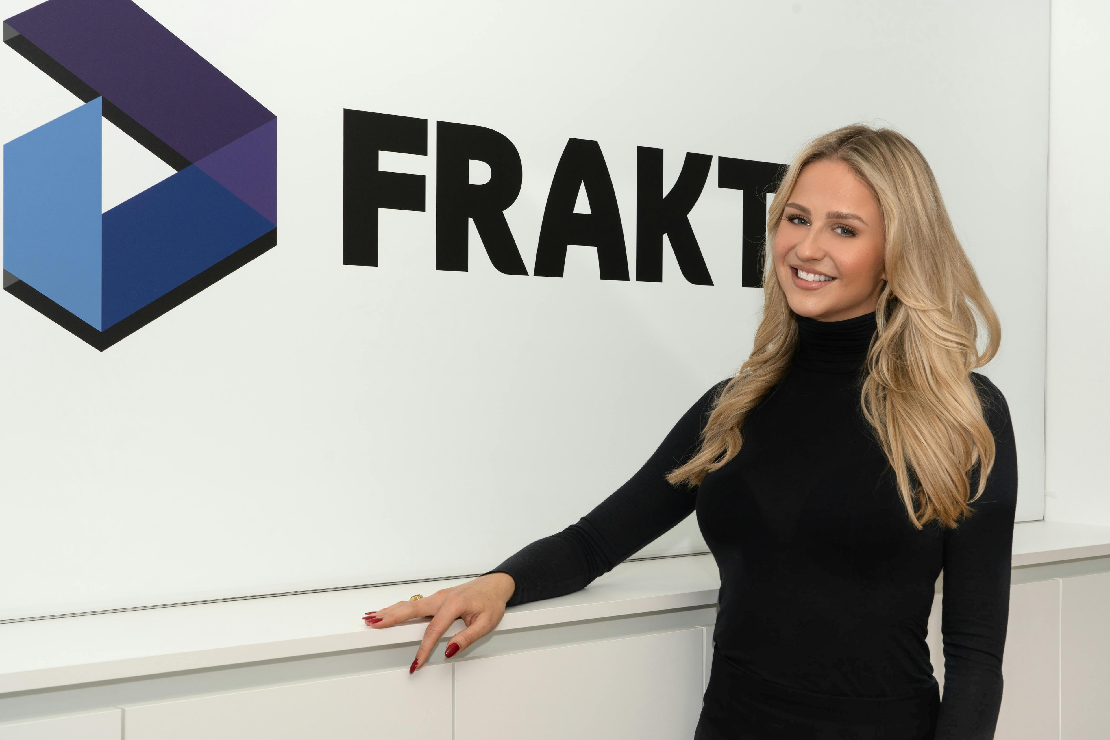 Isabell Lehmann, an associate partner at Fraktal Development, stands with a warm smile, leaning slightly against a counter. Her long blonde hair cascades over a stylish black turtleneck, complementing the professional ambiance. The prominent 'FRAKTAL' logo with its distinctive geometric design adorns the wall behind her, symbolizing the company's innovative edge.