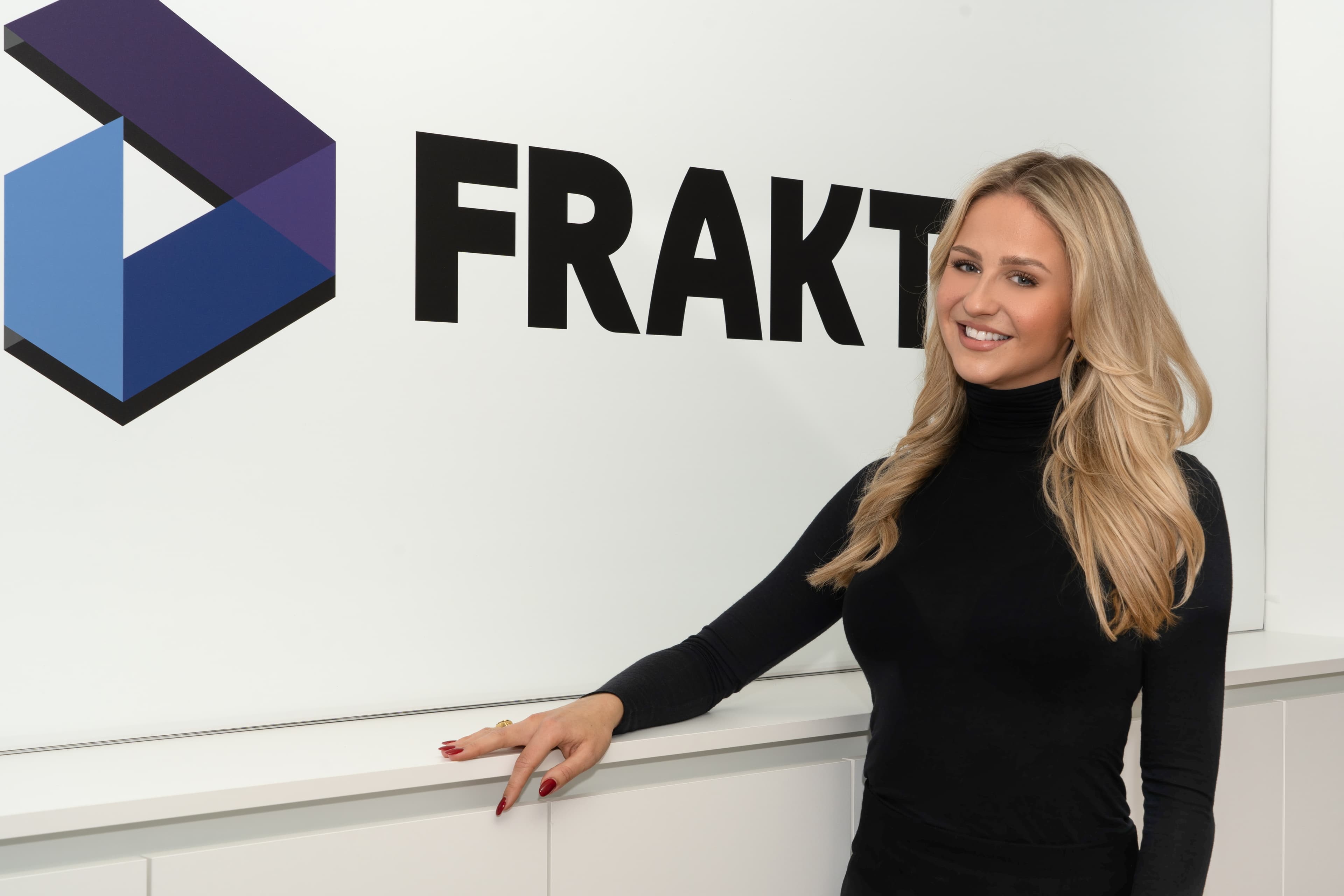 Isabell Lehmann, an associate director at Fraktal Development, stands with a warm smile, leaning slightly against a counter. Her long blonde hair cascades over a stylish black turtleneck, complementing the professional ambiance. The prominent 'FRAKTAL' logo with its distinctive geometric design adorns the wall behind her, symbolizing the company's innovative edge.