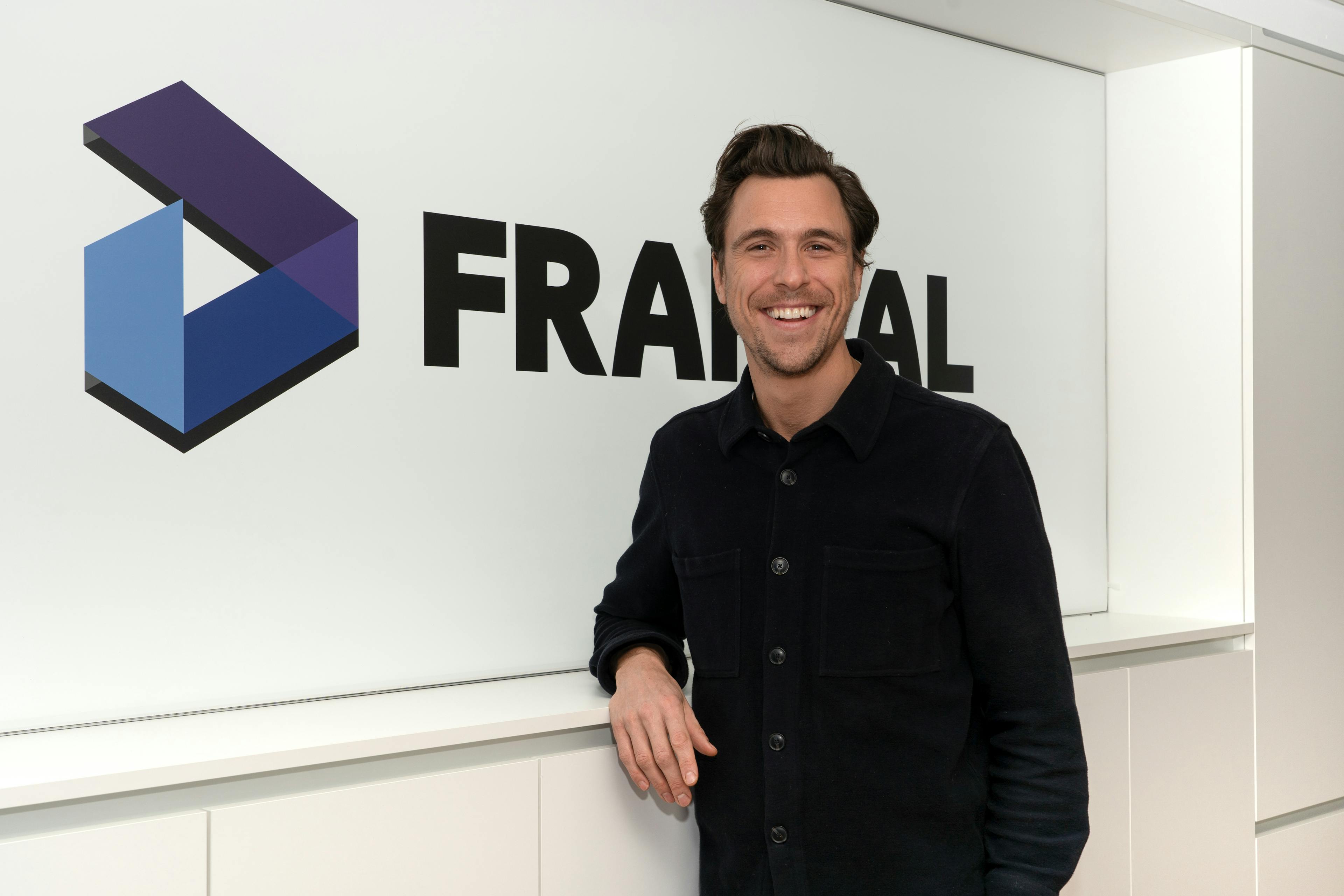 Kilian v. Dallwitz, the Press Spokesperson for Fraktal Development, presents a cheerful demeanor with a broad smile. He's dressed in a casual yet smart dark shirt, lending an approachable yet professional image. The 'FRAKTAL' logo looms prominently in the background, signifying the brand's strong presence.
