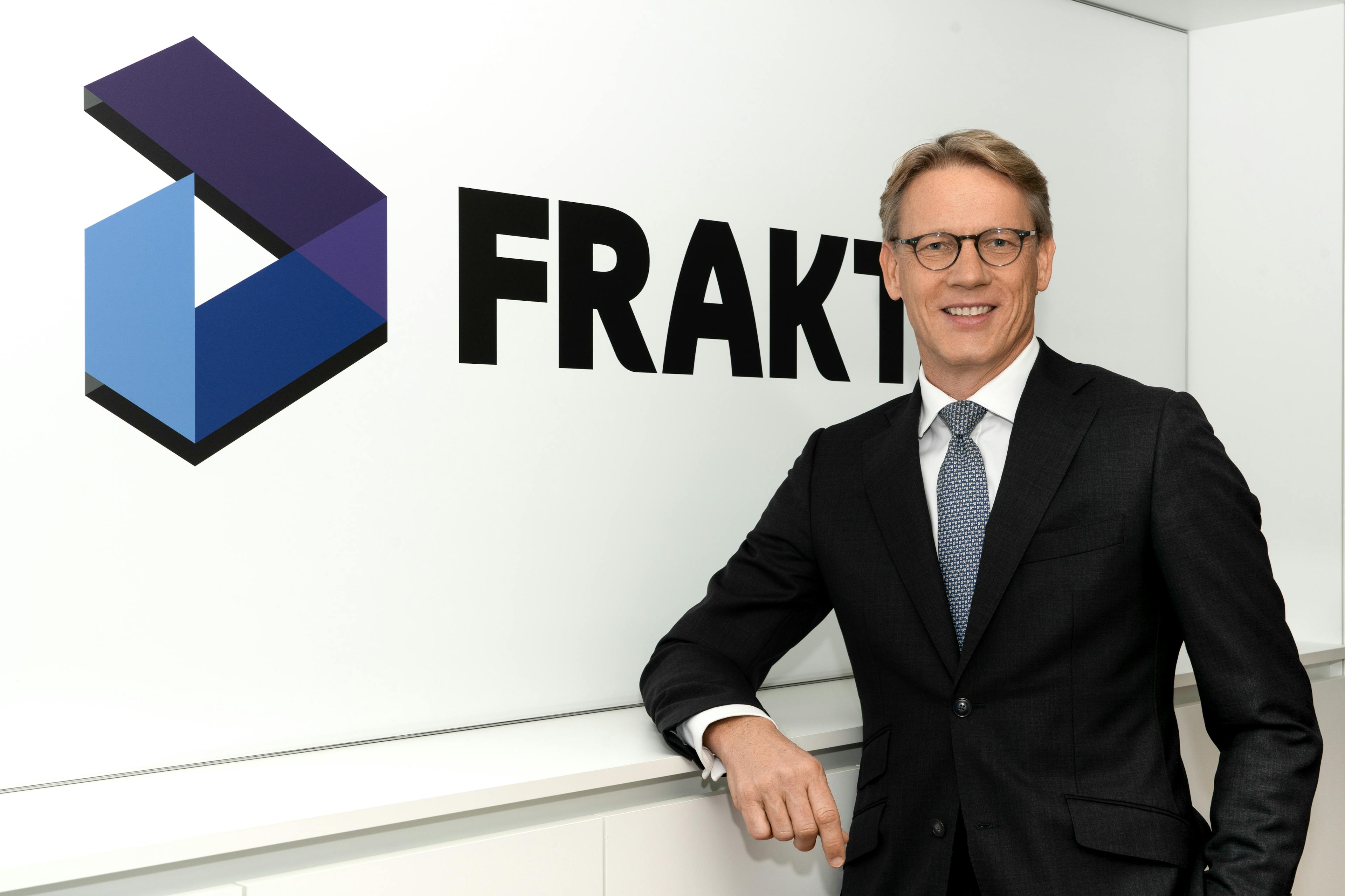 Portrait of Wolfgang Zauner, a partner at Fraktal Development, leaning casually with one hand on a white surface. He's wearing a sleek black suit, a crisp white shirt, and a patterned blue tie. Behind him, the bold 'FRAKTAL' logo with a stylized geometric symbol provides a dynamic and professional backdrop.
